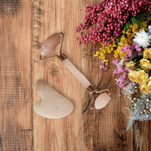 Gua Sha stone and facial roller on wooden table next to wildflowers.